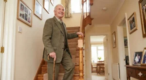 A senior standing safely at the bottom of a staircase, looking up confidently, holding a cane and a handrail